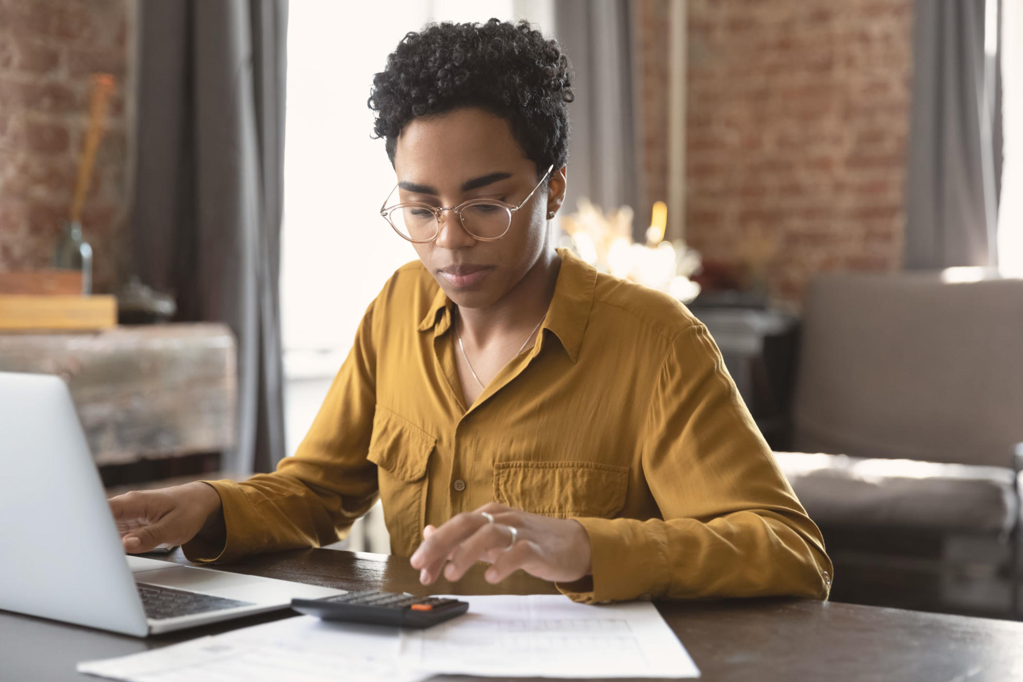 Focused millennial Black business woman calculating finance, money, using calculator, laptop computer at home workplace table calculating taxes