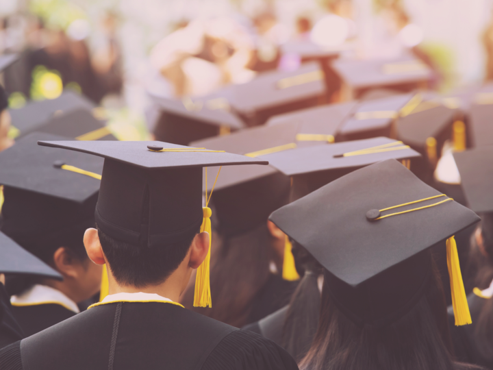 The backs of students in black graduation gowns and caps with yellow tassels.