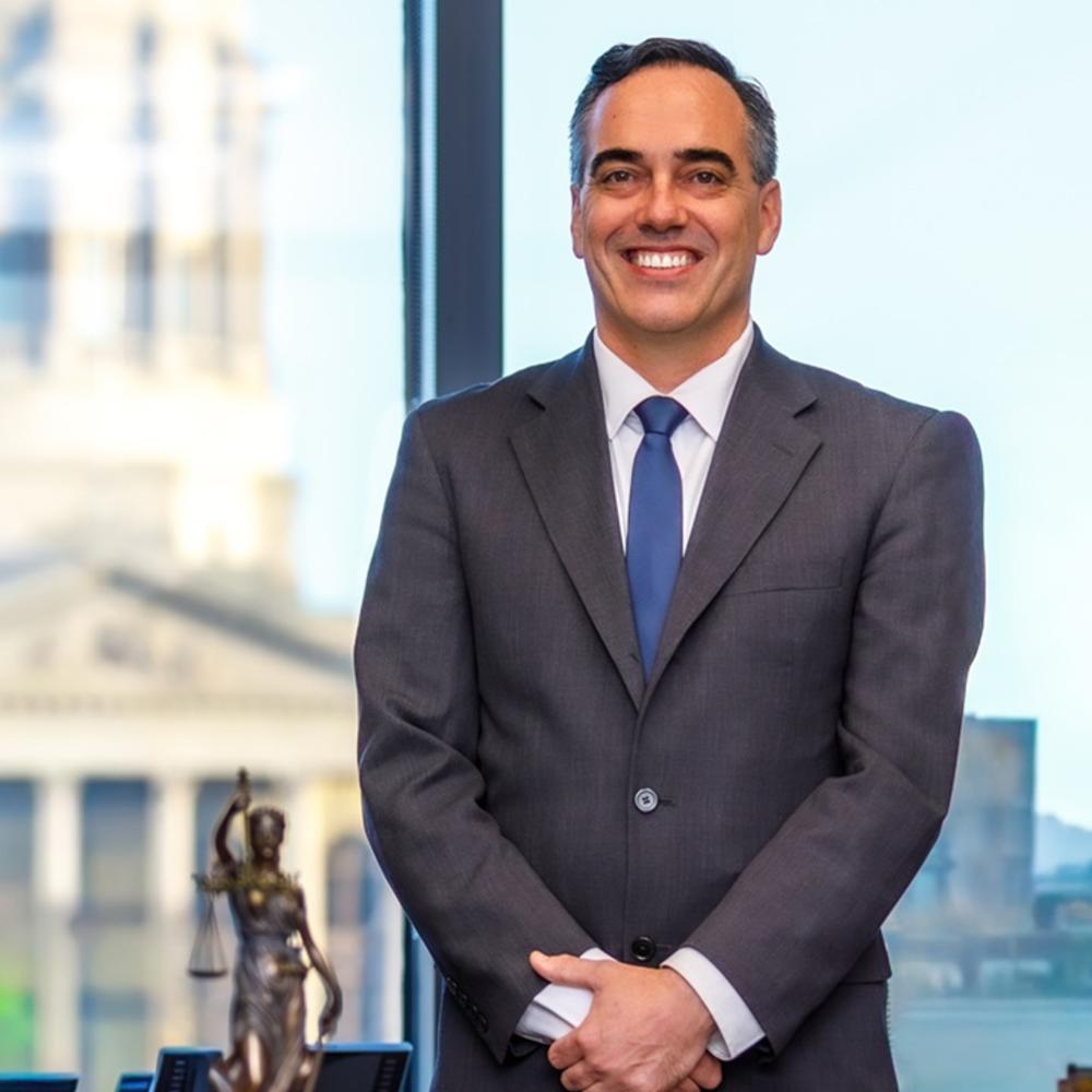 Commissioner Peter Hubbard wears a grey suit and blue tie and stands in front of large window